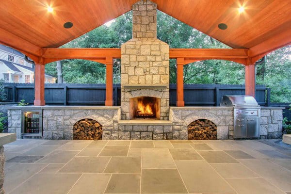 Outdoor patio with a stone fireplace, wooden ceiling, and a built-in grill next to stacked firewood