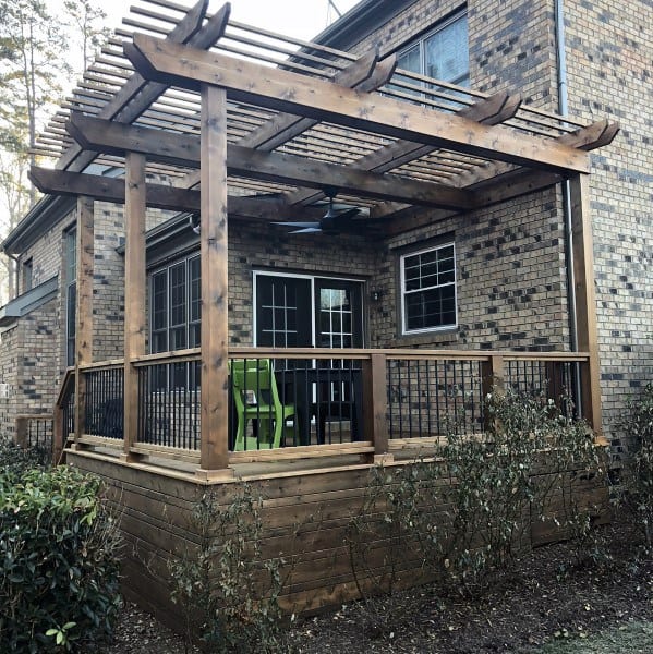 Wooden pergola with slatted roof, raised patio, railing, and a small seating area against a brick wall.