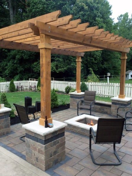 Wooden pergola with stone pillars, fire pit, and comfortable seating on a paved patio.