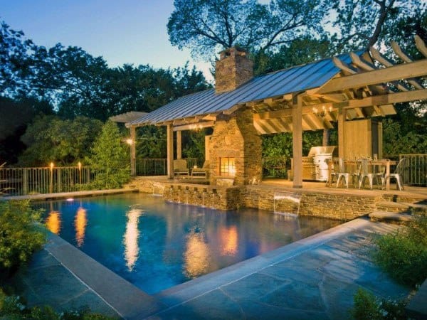 Stunning poolside pavilion with a stone fireplace, wooden pergola, outdoor dining area, and serene evening lighting.