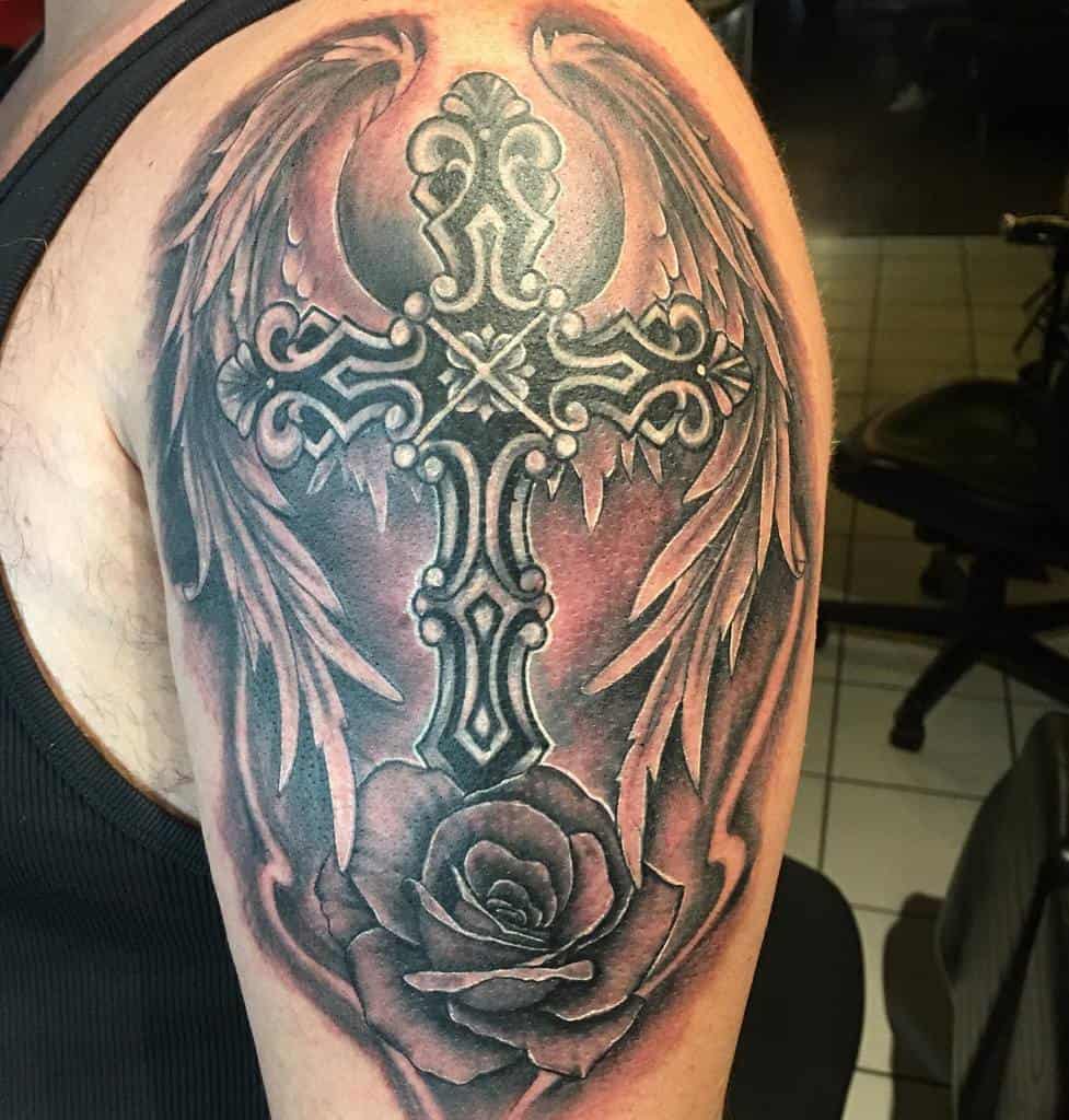 Tattoo of an ornate cross with angel wings and a rose below it with dark shading background