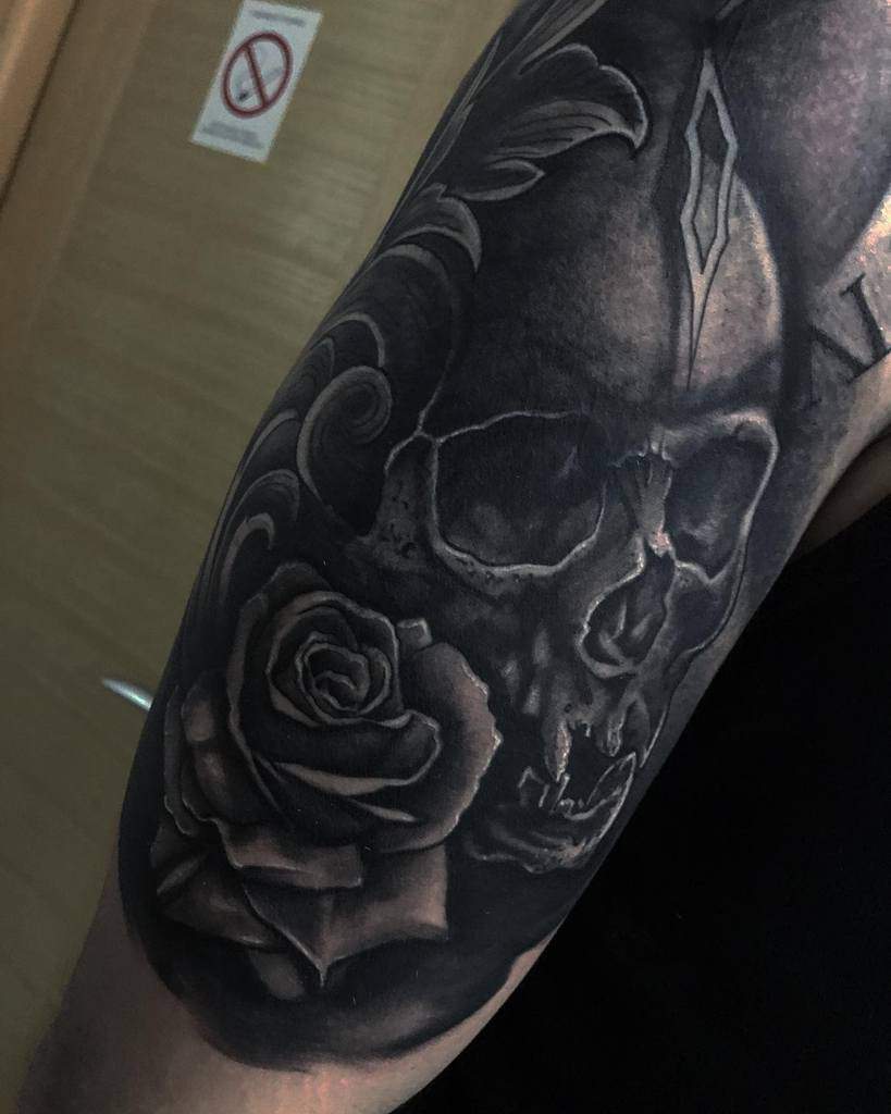 Dark and bold black and grey tattoo sleeve featuring a skull, a detailed rose, and ornamental elements with deep shading and contrast.