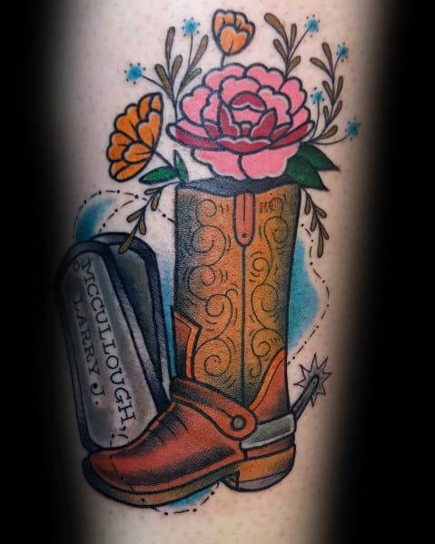 Tattoo of a cowboy boot with floral details, dog tags, and a spur, symbolizing Western heritage, remembrance, and personal tribute.