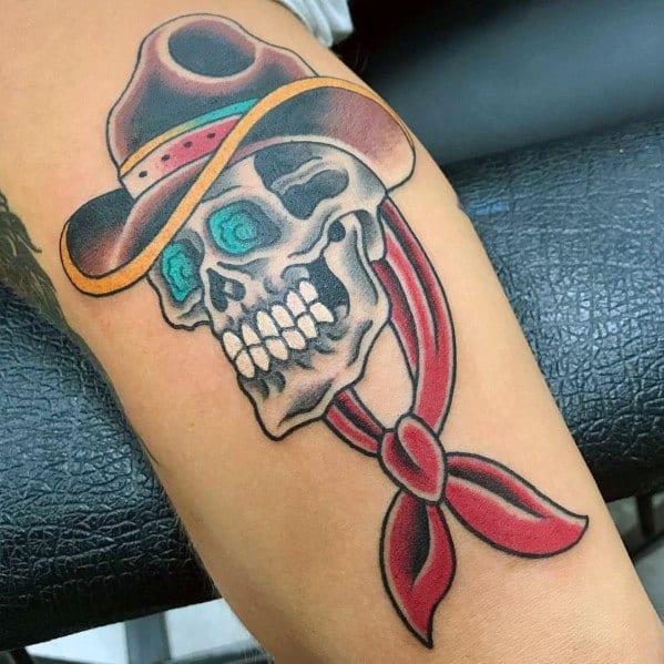 Tattoo of a cowboy skull with glowing blue eyes, wearing a brown hat and red bandana, done in a bold traditional style with vibrant colors.