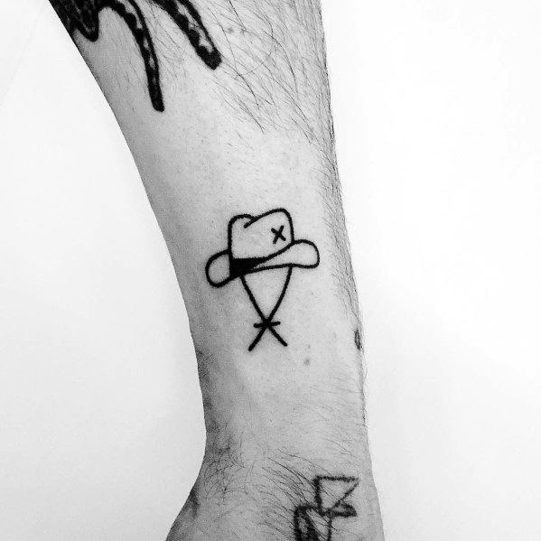 Minimalist tattoo of a cowboy hat with a small "X" for an eye and crossed lines beneath, creating a simple and abstract Western design.