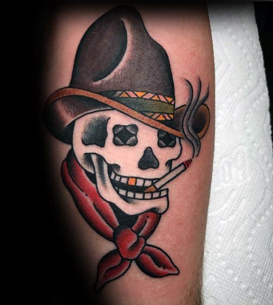 Tattoo of a grinning cowboy skull with star-shaped eyes, wearing a brown hat and red bandana, smoking a cigarette with bold shading.