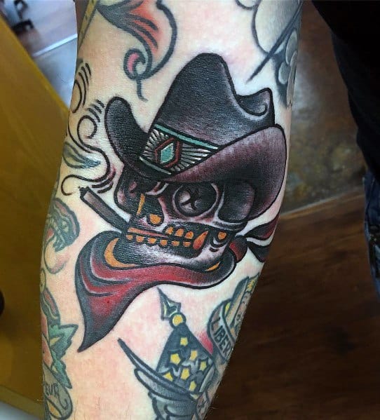 Tattoo of a cowboy skull with a black hat, red bandana, and a cigarette, featuring bold colors, shading, and a rugged Western style.