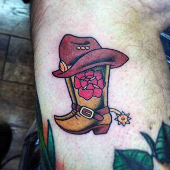Tattoo of a cowboy boot with a floral design, wearing a brown hat, and featuring a detailed spur, done in a bold and colorful Western style.