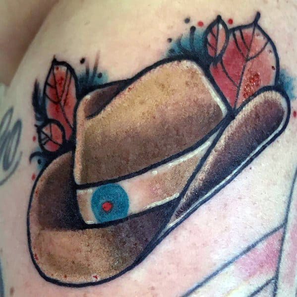 Tattoo of a brown cowboy hat with a decorative band, featuring red leaves and blue accents, done in a bold Western style with shading details.