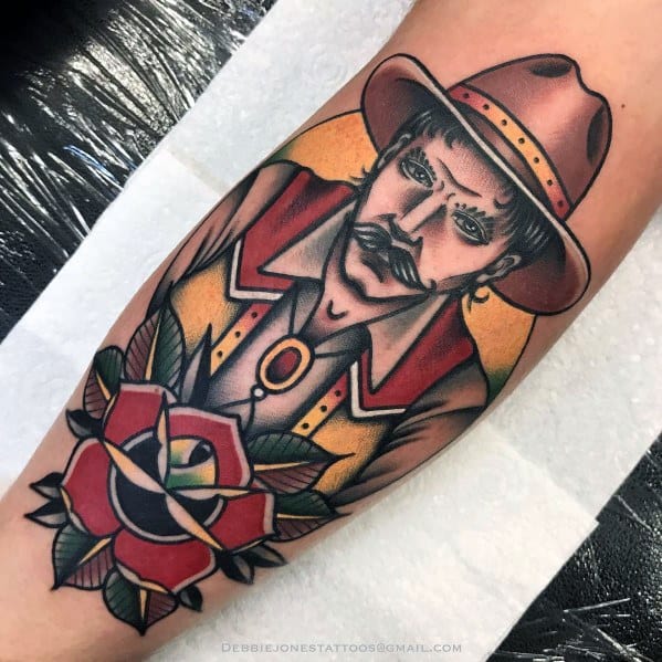 Tattoo of a cowboy with a mustache wearing a brown hat and vest, set against a sunset background, with a bold red rose in the foreground.