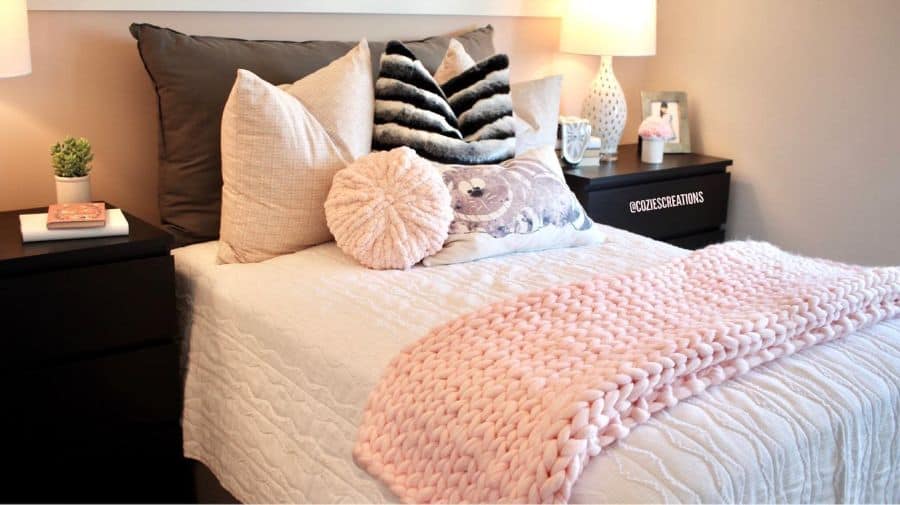 Cozy bedroom with plush pillows, a pink knitted blanket, bedside lamps, and decorative accessories
