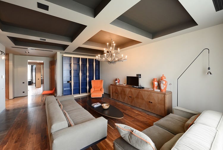 A cozy living room featuring a coffered ceiling, stylish modern furniture, bold orange accents, and a chandelier, creating a comfortable yet sophisticated atmosphere