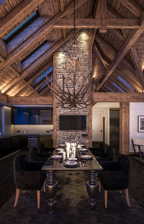 Rustic dining room with wooden beams, antler chandelier on the ceiling, stone fireplace, set table, and plush chairs