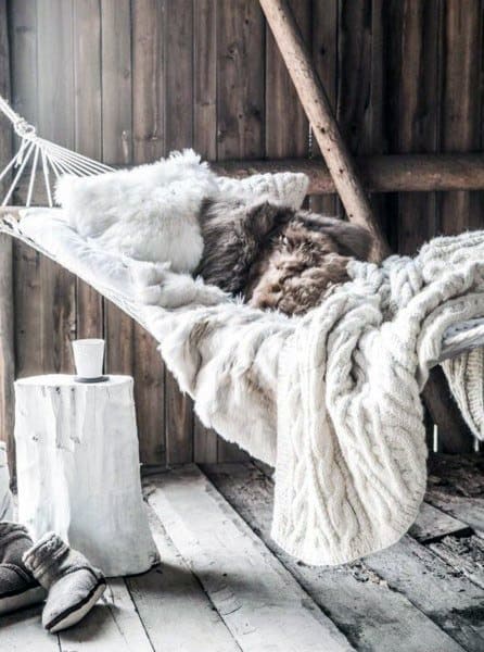 Cozy hammock in a rustic wooden room with fluffy blankets, pillows, and a small white table.