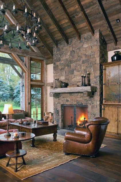 Rustic living room with tall stone fireplace, leather armchair, wooden beams, and large windows.