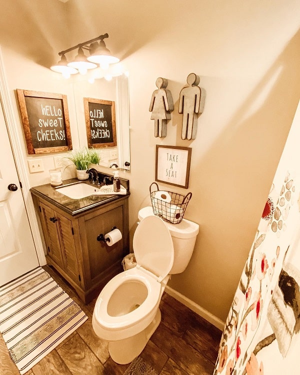 A cozy farmhouse bathroom with humorous decor, dual mirrors, a vanity, and toilet. Signs read "Hello Sweet Cheeks" and "Take a Seat