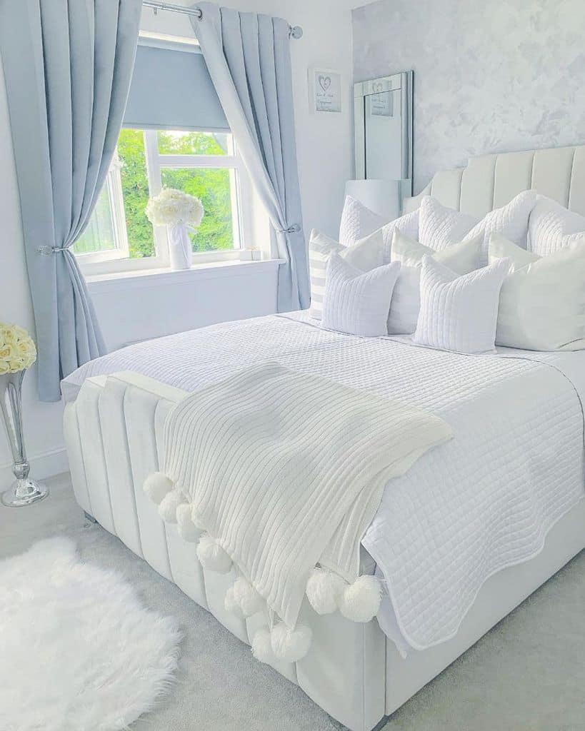 Bright bedroom with a crisp white bed, fluffy pillows, gray curtains, and a window letting in natural light
