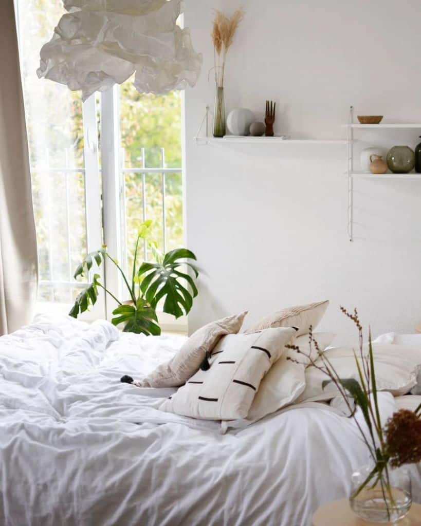 Cozy bedroom with a white bed, plush pillows, and a plant by the window; shelves display decor and a pendant light hangs overhead