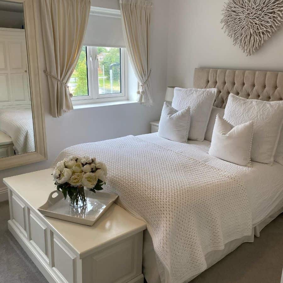 Cozy bedroom with a neatly made bed, white decor, a window, and a floral arrangement on a white chest