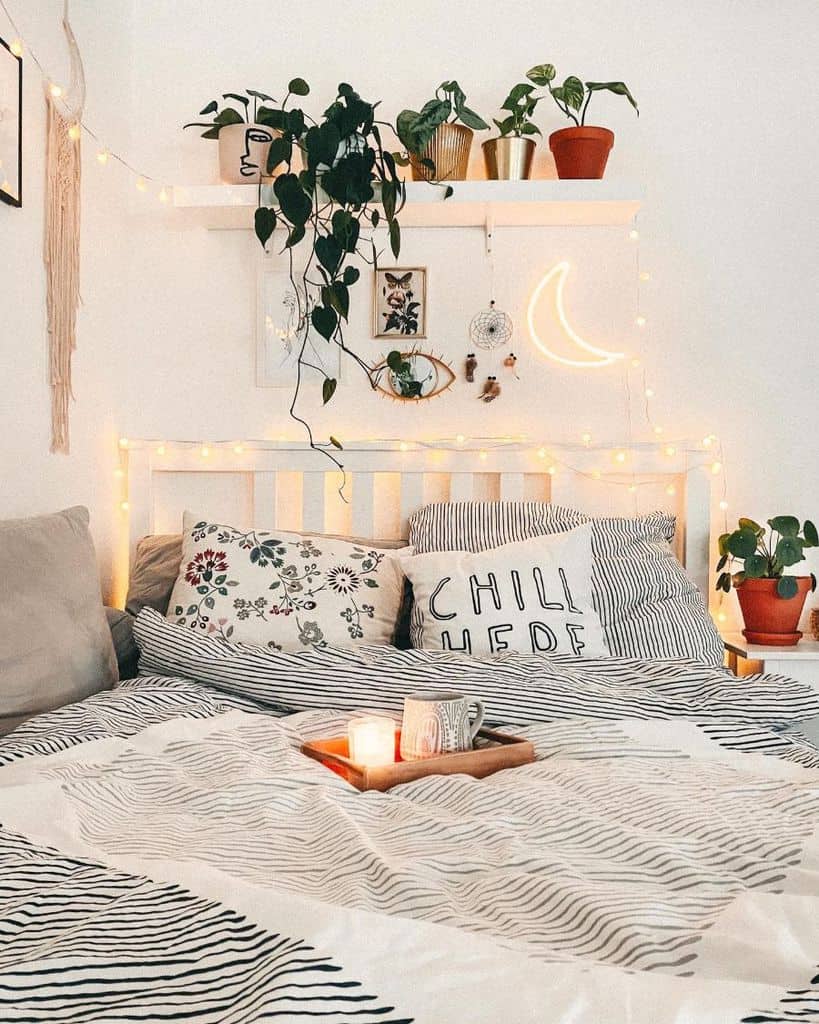 Cozy white bedroom with string lights, plants, and cushions. A candle and small tray rest on the blanket