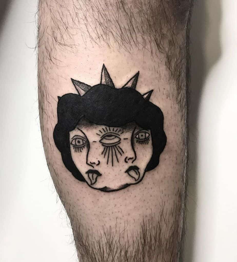 Tattoo of a surreal two-faced figure with an all-seeing eye on the forehead, a crown-like headpiece, and bold black shading, inked on the leg.