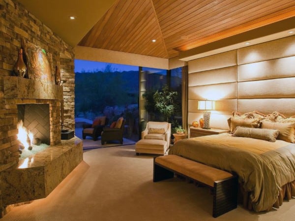 Luxury cabin bedroom with wood ceiling, stone fireplace, and upholstered headboard.
