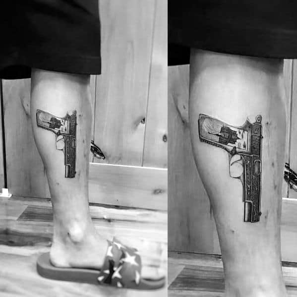 Realistic black and gray tattoo of an engraved 1911 pistol on the calf, featuring intricate details and shading for a bold three-dimensional look.