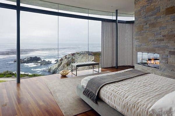 Modern bedroom with stone fireplace, glass walls, and stunning ocean cliff views.