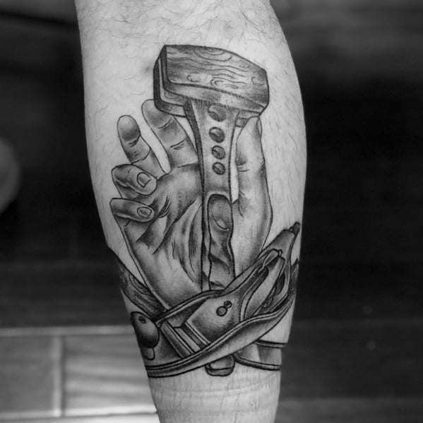 Black and gray tattoo of a hand gripping a mallet, wrapped in a measuring tape, symbolizing strength, craftsmanship, and dedication to woodworking.