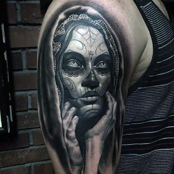 La Catrina tattoo on upper arm showing a woman with sugar skull makeup, lace veil, and hands framing her face in dramatic shading.