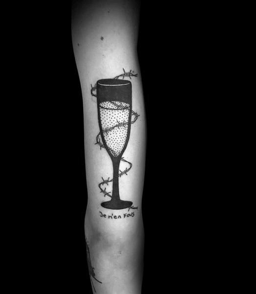 Arm tattoo of a champagne glass with bubbles, surrounded by thorns, against a black background.