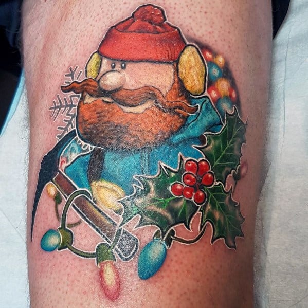 Christmas tattoo of Yukon Cornelius from *Rudolph the Red-Nosed Reindeer*, holding an ice pick, surrounded by holly, snowflakes, and festive lights.