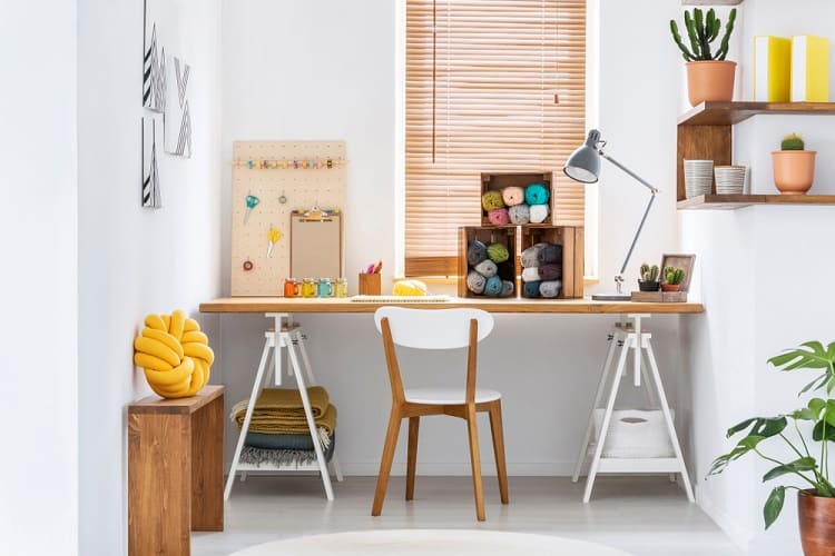 Bright craft room with a wooden desk, chair, yarn, potted plants, and a lamp under a window with blinds
