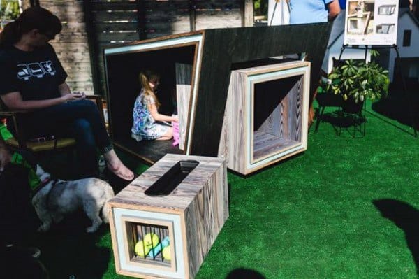 Modern, creatively designed dog house with angular wooden structures, featuring a playful space for both a dog and a child, surrounded by lush green grass