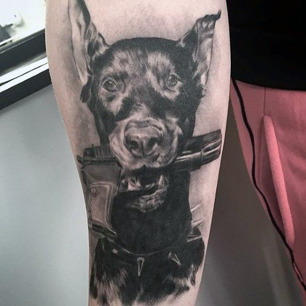 Realistic black and grey Doberman tattoo showing the dog gripping a rifle in its mouth, with sharp detailing and a fierce expression.
