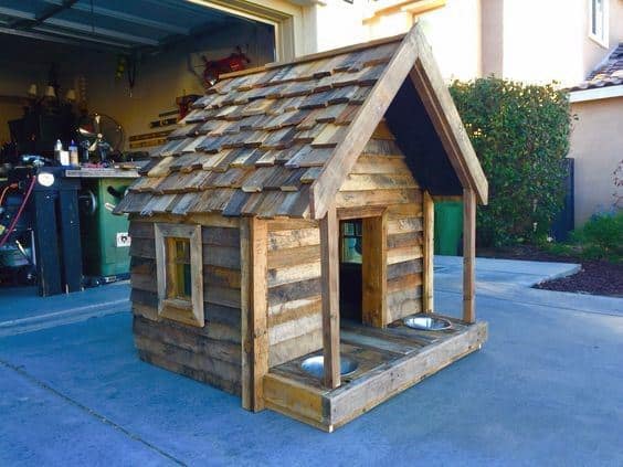 A small wooden doghouse with a shingled roof, two metal bowls in front
