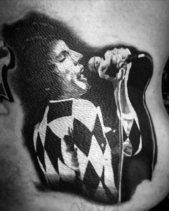 Black and grey Freddie Mercury tattoo in harlequin jumpsuit, passionately singing into a microphone with dramatic lighting.