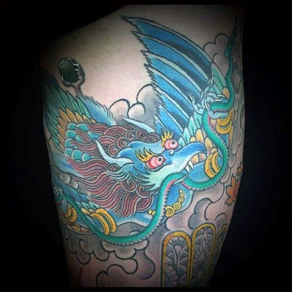 Colorful Garuda tattoo on the thigh featuring a fierce blue bird-like creature with vibrant wings, clouds, and flowing ornamental details.