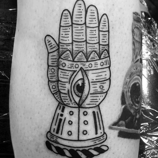 Black ink tattoo of a stylized armored hand with an eye in the palm, featuring geometric patterns and bold linework.