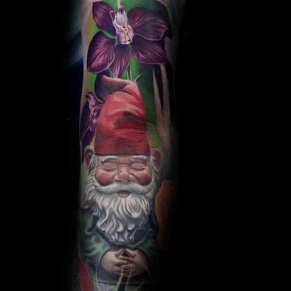 Gnome tattoo with a red hat and white beard, sitting calmly with hands clasped, surrounded by greenery and a vibrant purple flower.