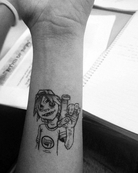Simple black line tattoo of 2D from Gorillaz on the wrist, smiling and holding up his fingers in a peace sign with a casual look.