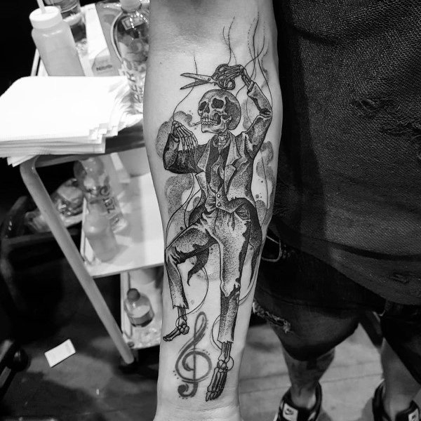 Black and grey forearm tattoo featuring a skeletal puppet master in a suit, controlling strings, with a treble clef symbol for a gothic aesthetic.