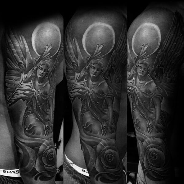 A detailed black and grey tattoo on the upper arm featuring an angel with large wings, a halo, and roses, positioned in a flowing and dynamic design.