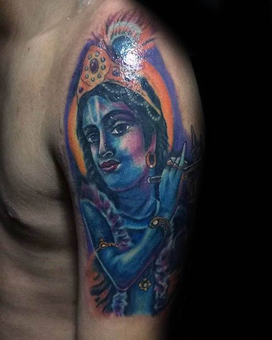 Colorful upper arm Krishna tattoo featuring a radiant blue-skinned Krishna with a halo, playing the flute and wearing a peacock-feathered crown.