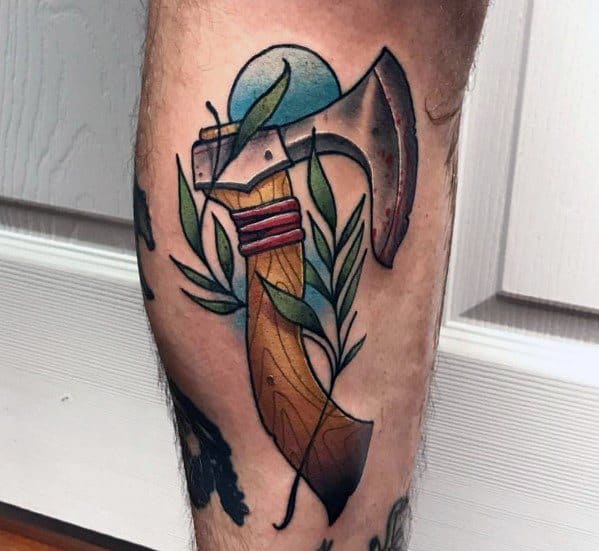Colorful hatchet tattoo on the leg featuring a weathered blade, wooden handle, red binding, and green leaves, with a blue circular background.