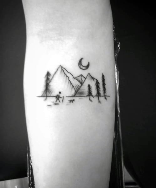 Minimalist hiking tattoo featuring mountains, trees, a crescent moon, and a hiker with a dog, symbolizing adventure and nature.