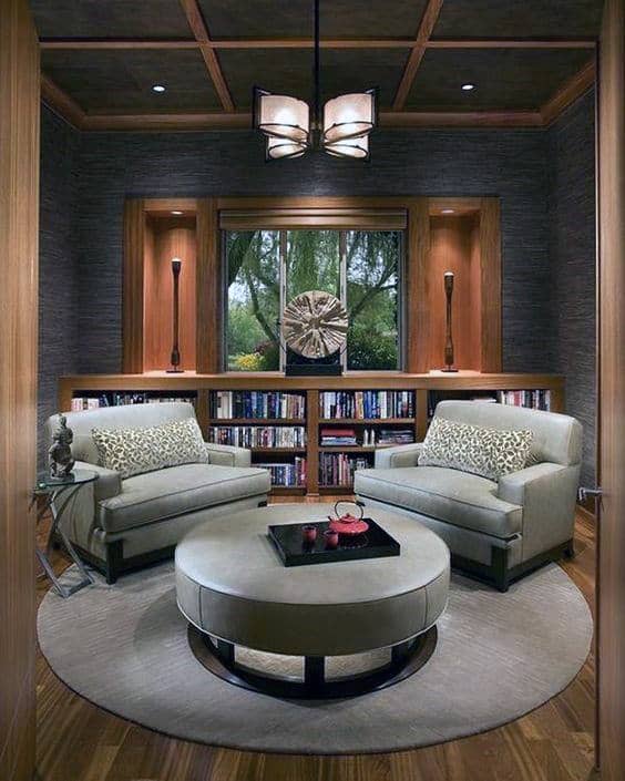 Cozy living room with two armchairs, library-style bookshelves, a round ottoman, and a large window overlooking greenery
