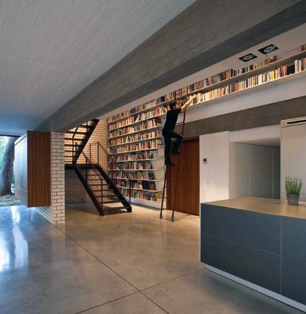 Person on ladder reaching for books on a tall bookshelf in a modern library-themed home with concrete and brick elements