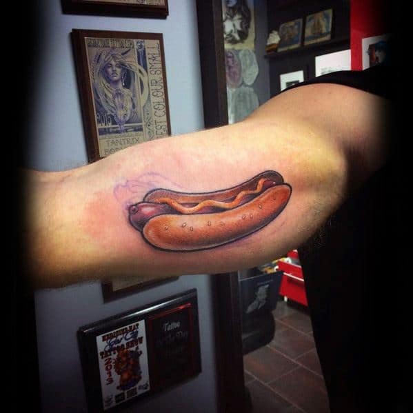 Hot dog tattoo on the forearm featuring a sausage with mustard in a soft bun, set against a tattoo studio background.