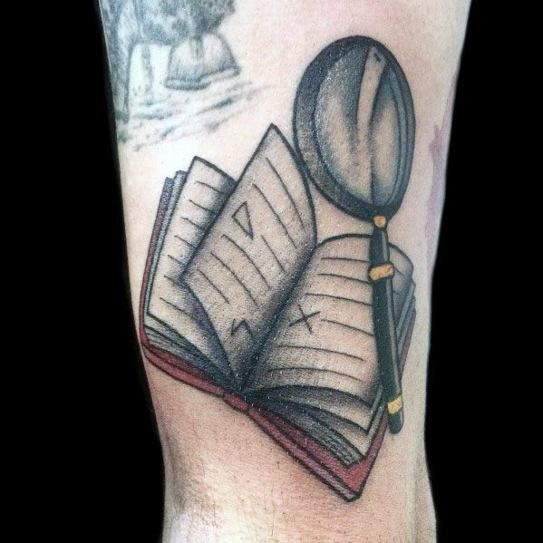 Tattoo of an open book with a magnifying glass, featuring bold outlines and subtle shading for a classic, old-school design.